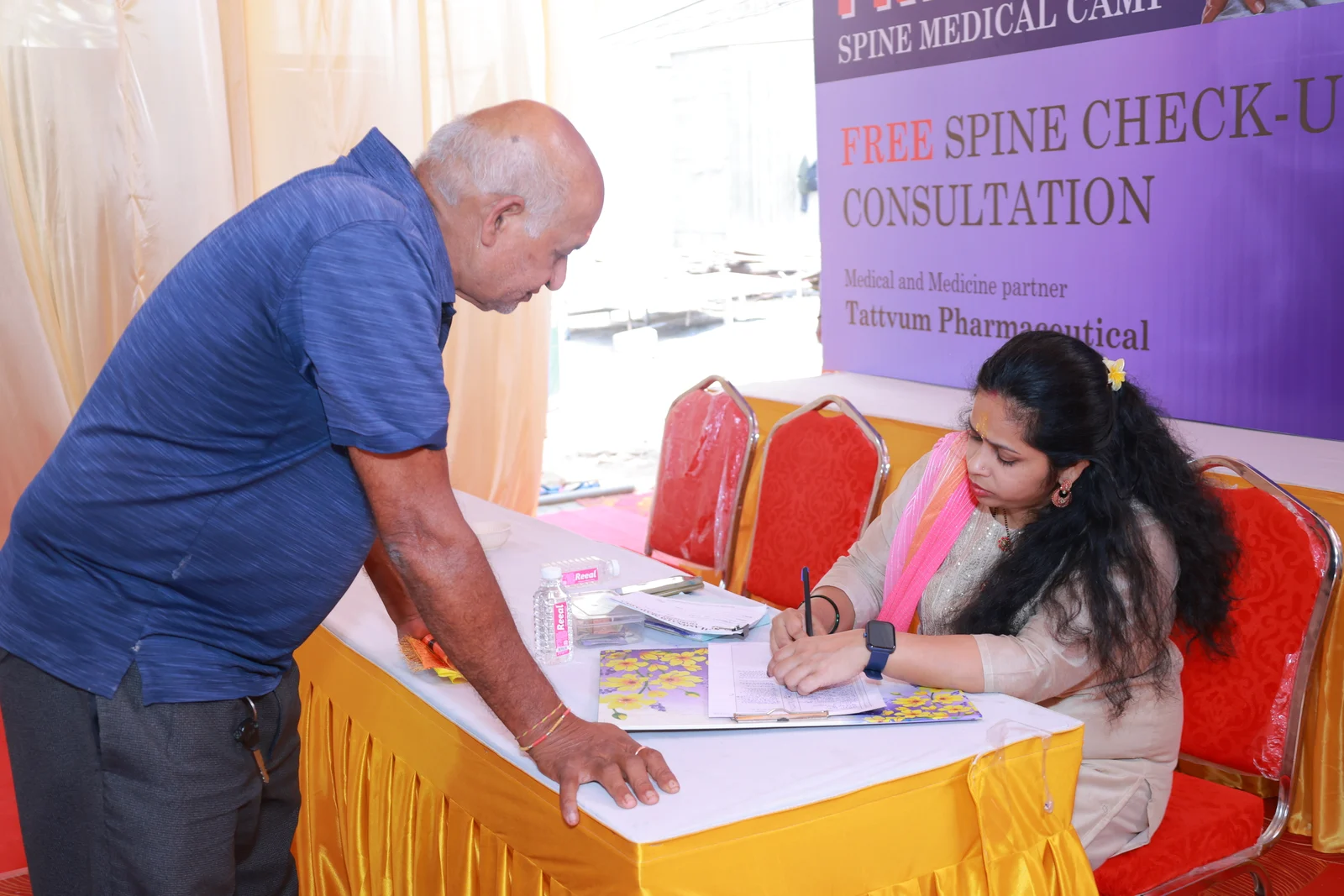 A man leaning across the spine consultation desk while a seated woman writes on a form