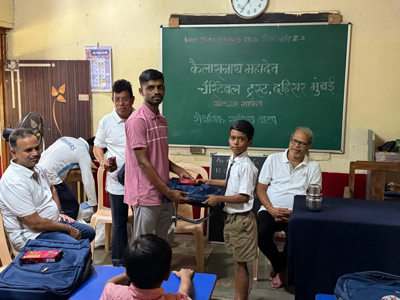A boy and a presenter holding a school bag together in front of a classroom blackboard